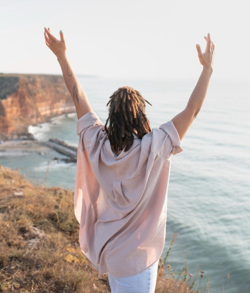 woman holds her arms up triumphantly by the seaside, she understands self-pleasure